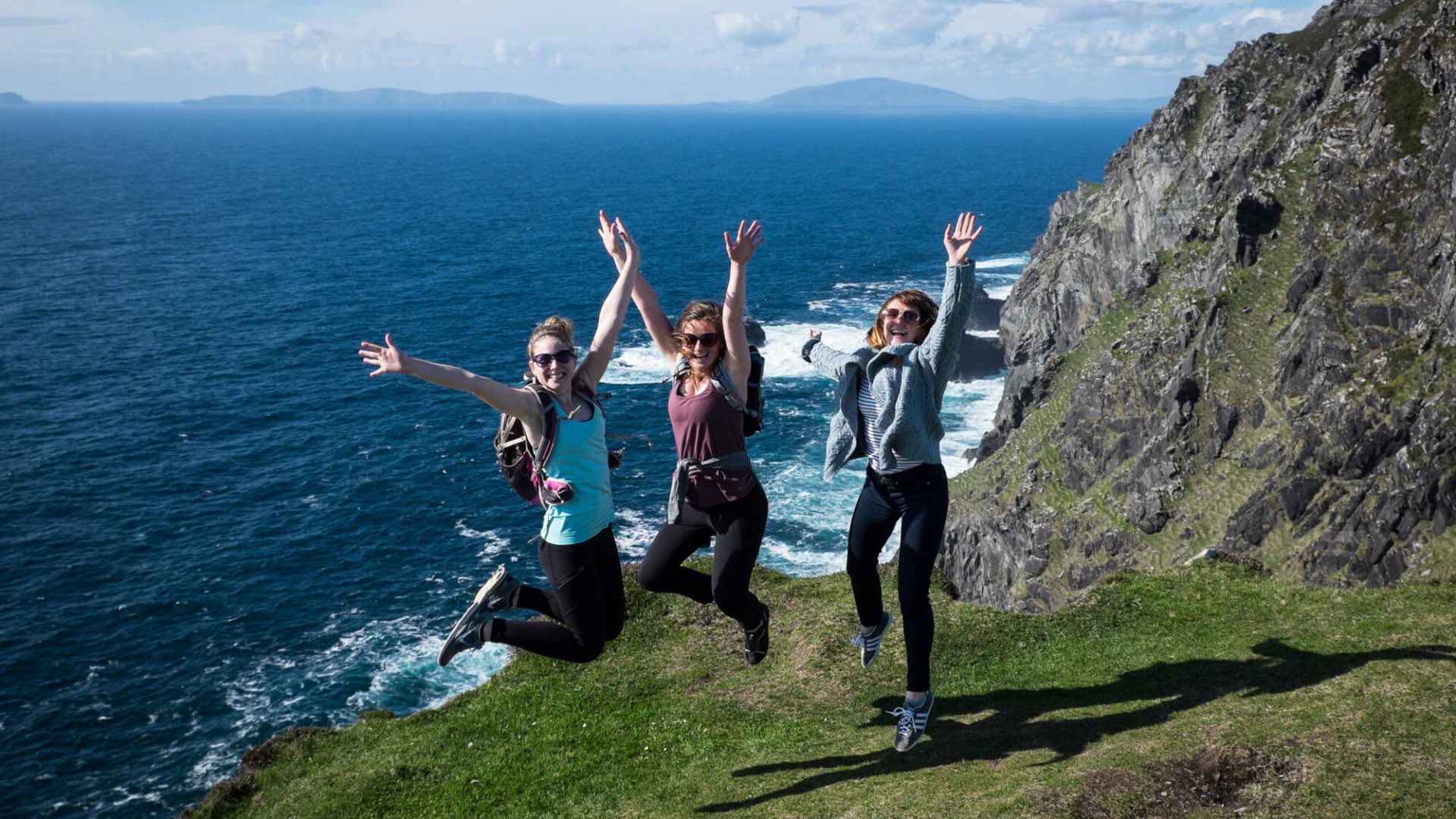 Three female tour guests wearing hiking gear and jumping in the air on Bray Head, Valentia Island, Ireland