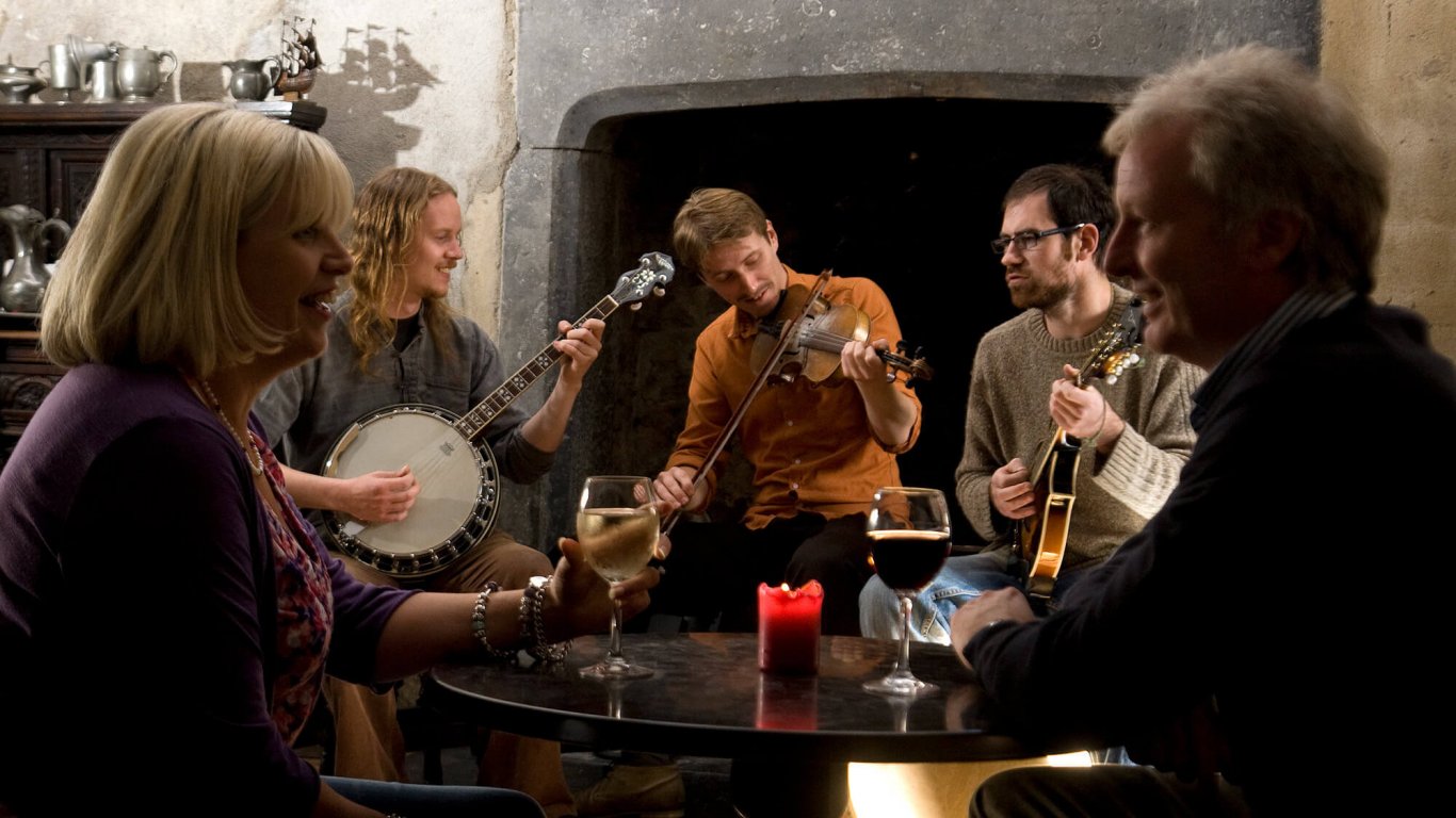A middle-aged couple face each other over a table while three younger traditional Irish musicians play in the background