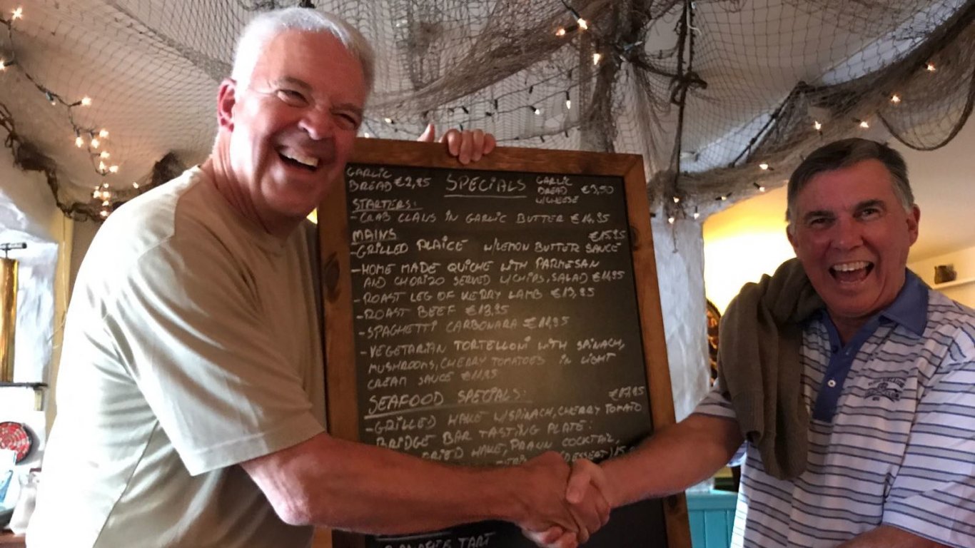 Two smiling Vagabond passengers shake hands in front of a specials board at The Moorings restaurant and bar in Portmagee on the Ring of Kerry, Ireland