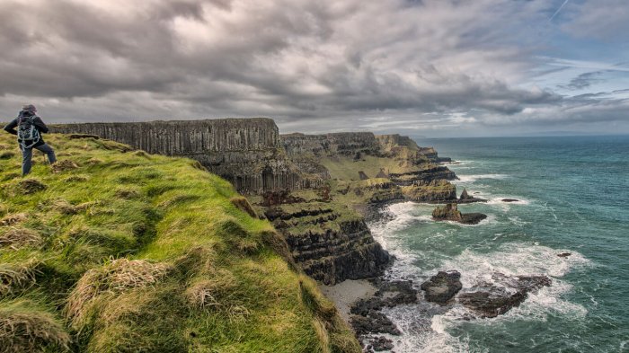 A lone hiker looks out over a scenic coastal scene of cliffs, moody clouds and big waves