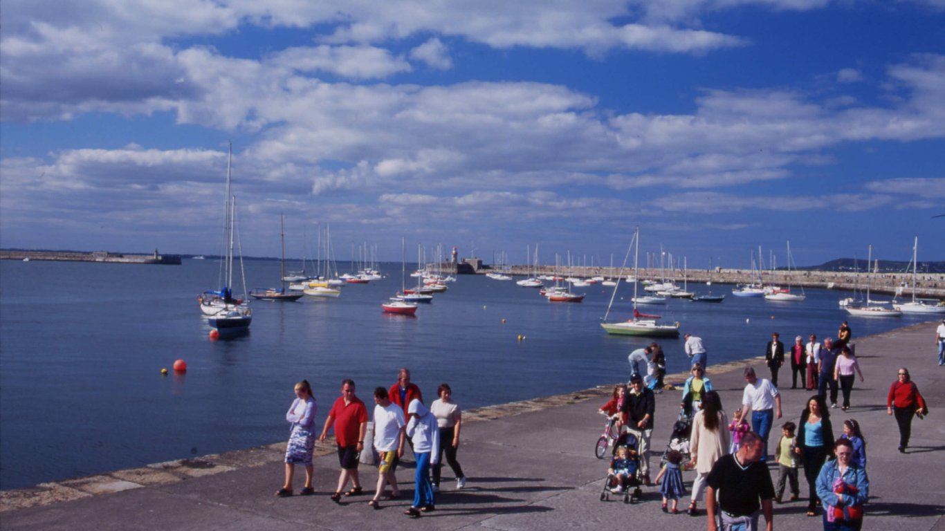 Dun Laoghaire pier with a view of all the sailing boats out in the sunshine and people walking on the pier A view of Dun Laoghaire Harbour with boats sailing in the sunshine and people walking on the pier